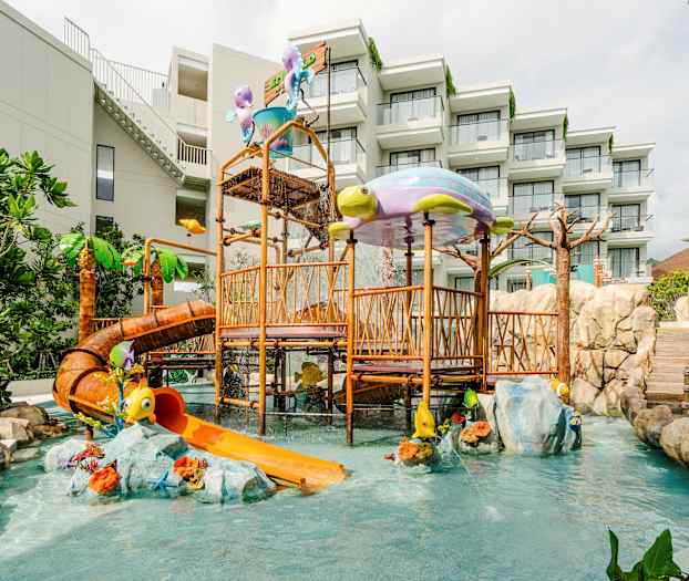 Leikkipaikka vesiliukumäellä ja leikkivälineillä, ympärillä vihreitä kasveja ja kivimuureja. Hotellirakennus taustalla. (Tekoälyn luoma teksti)