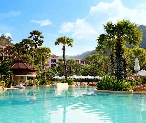 Uima-allas, jota ympäröivät korkeat palmut, aurinkotuolit varjoineen ja rakennukset vihreyden keskellä. (Tekoälyn luoma teksti)