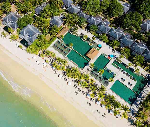 Ilmakuva rannasta, jossa on aurinkovarjoja, palmuja, uima-allasalue ja rakennuksia. (Tekoälyn luoma teksti)