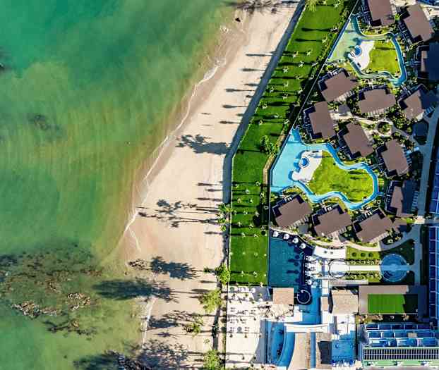 Ilmakuva rannasta ja hotellialueesta, jossa on uima-altaita ja rakennuksia. (Tekoälyn luoma teksti)