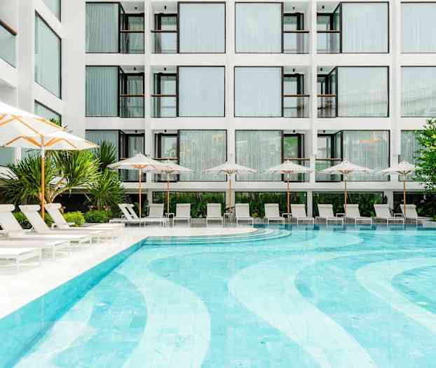 Moderni rakennus, jossa on suuret ikkunat, näkyy taustalla, edessä uima-allas, jota ympäröivät aurinkotuolit ja aurinkovarjot. Kasvit ja vihreä ympäristö rikastuttavat aluetta. (Tekoälyn luoma teksti)