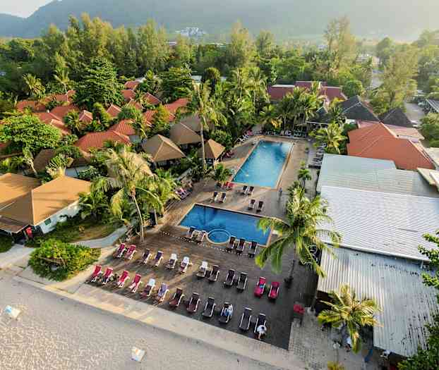 Ilmakuva lomakeskuksesta, jossa on punaiset kattot, kaksi allasaluetta ja aurinkotuoleja lähellä hiekkarantaa. (Tekoälyn luoma teksti)