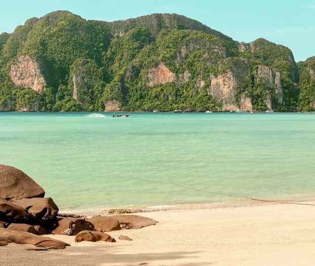 Hiekkaranta, jossa etualalla on kallioita, kirkkaan turkoosi vesi ja taustalla vihreitä vuoria. (Tekoälyn luoma teksti)