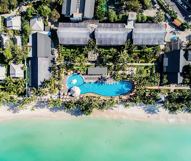 Ilmakuva lomakeskuksesta, jossa on uima-allas, trooppisten puiden ympäröimänä ja lähellä kirkasvetistä rantaa. (Tekoälyn luoma teksti)