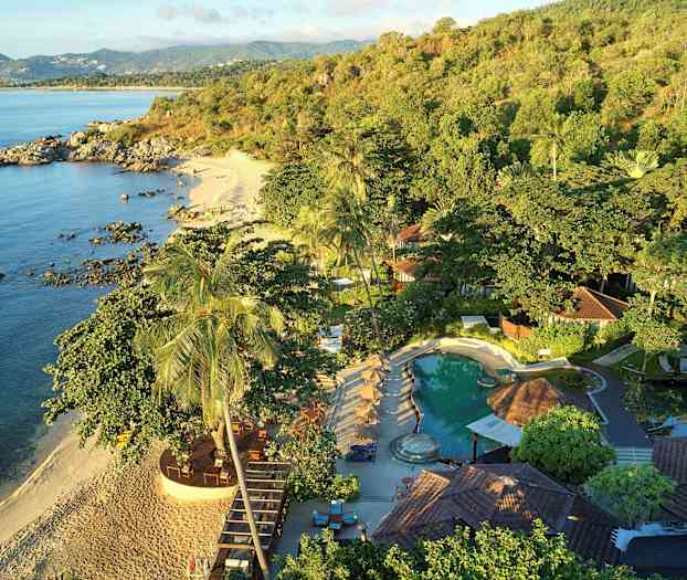 Kuva, jossa on trooppinen ranta, jota ympäröi vehmas kasvillisuus, ja etualalla on uima-allas ja rakennuksia. (Tekoälyn luoma teksti)