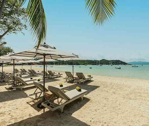 Hiekkaranta, jossa aurinkotuoleja ja varjoja; taustalla meri ja veneitä. (Tekoälyn luoma teksti)