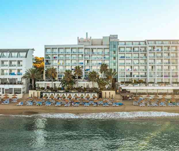 Kuva suuresta valkoisesta hotellirakennuksesta aurinkoisella rannalla, jossa on aurinkotuoleja ja aurinkovarjoja. (Tekoälyn luoma teksti)