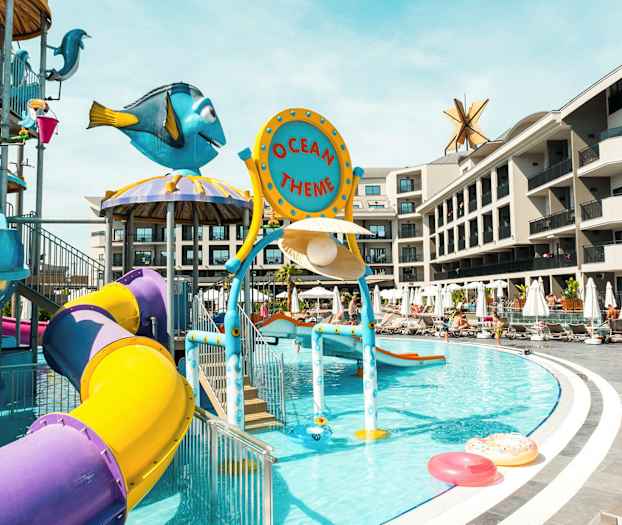 Kuva vesileikkipaikasta, jossa on värikkäitä liukumäkiä ja teemakoristelu altaalla. Taustalla näkyy hotellirakennuksia. (Tekoälyn luoma teksti)