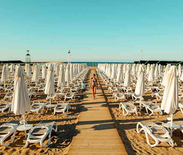 Nainen bikineissä kävelee puupolkua pitkin rantatuolien ja aurinkovarjojen välissä rannalla. (Tekoälyn luoma teksti)