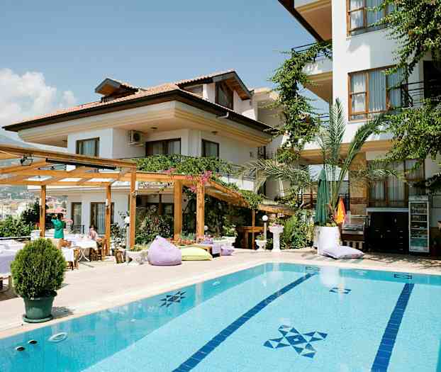 Valkoisella julkisivulla varustettu rakennus, jota ympäröivät vihreät kasvit, ja etualalla on uima-allas. (Tekoälyn luoma teksti)