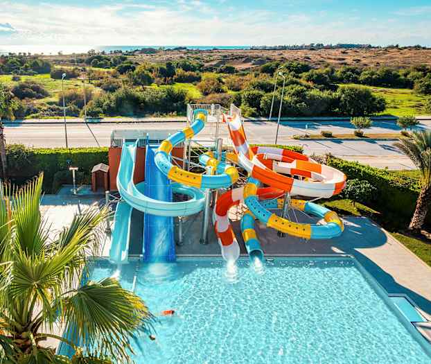 Kuvassa on vesipuisto, jossa on useita värikkäitä vesiliukumäkiä ja uima-allas, ympäröitynä vehreällä maisemalla. (Tekoälyn luoma teksti)