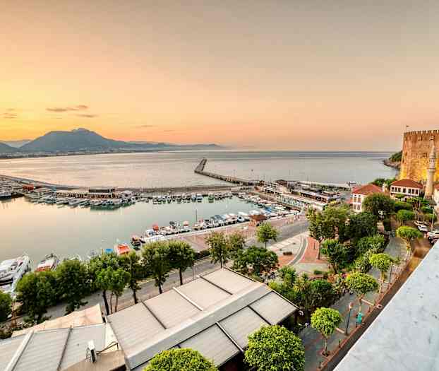 Kuvassa näkyy satama, jossa veneitä ja rakennuksia meren äärellä auringonlaskun aikaan. Oikealla näkyy linna. (Tekoälyn luoma teksti)