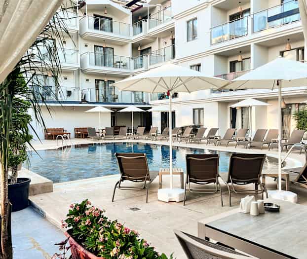 Kuva näyttää hotellin uima-altaan, jota ympäröivät aurinkotuolit, aurinkovarjot ja parvekkeet taustalla. (Tekoälyn luoma teksti)