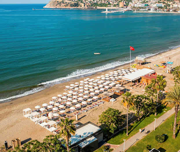 Ilmakuva rannasta, jossa on aurinkotuoleja ja aurinkovarjoja, turkoosi meri ja rakennuksia taustalla. (Tekoälyn luoma teksti)