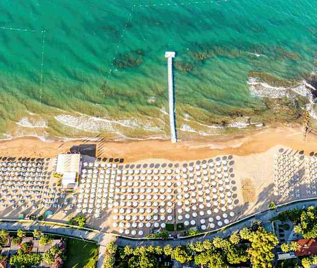 Ilmakuva rannasta, jossa on aurinkovarjoja ja aurinkotuoleja sekä laituri, joka ulottuu turkoosiin veteen. (Tekoälyn luoma teksti)