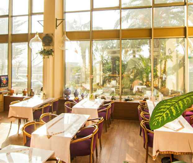 Kuva vaaleasta kahvilasta, jossa on suuret lasi-ikkunat, koristeltu pöydillä ja tuoleilla, joissa on violetit pehmusteet. (Tekoälyn luoma teksti)