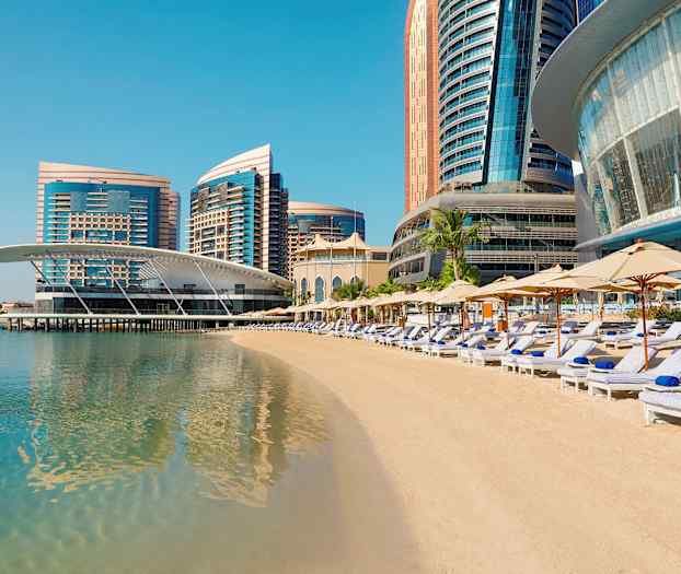 Ranta, jossa aurinkotuoleja ja varjoja, modernien tornitalojen edessä meren rannalla. (Tekoälyn luoma teksti)