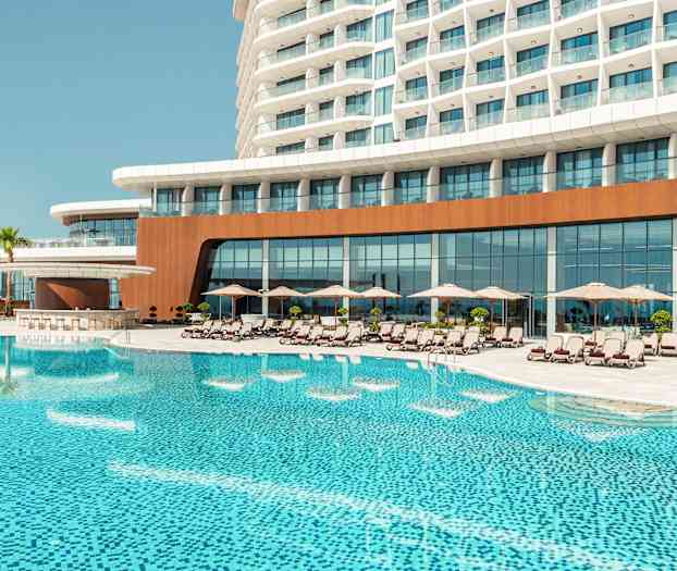 Hotellirakennus, jonka edessä on suuri uima-allas, ympärillä aurinkotuoleja ja aurinkovarjoja. (Tekoälyn luoma teksti)