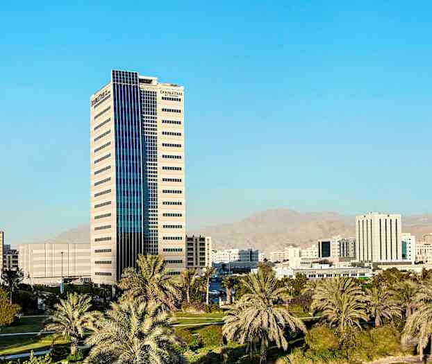 Kaupunkinäkymä, jossa korkea rakennus on ympäröity palmujen ja muiden rakennusten kanssa taustalla. (Tekoälyn luoma teksti)