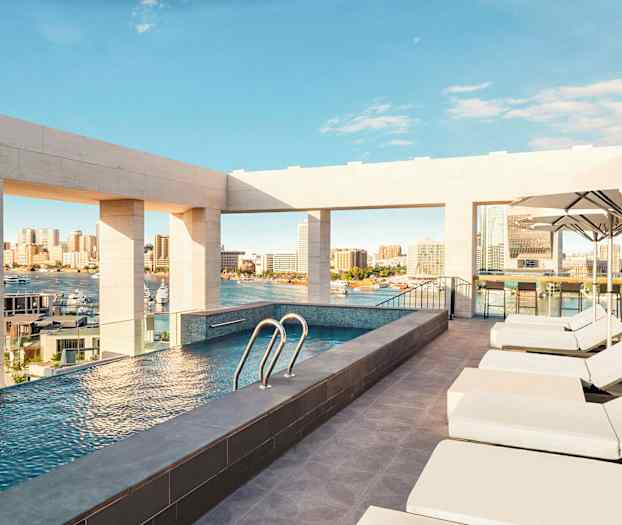 Kattoikkunalla varustettu uima-allas tarjoaa näkymät kaupunkiin ja satamaan, ympärillä aurinkotuoleja ja varjoja. (Tekoälyn luoma teksti)