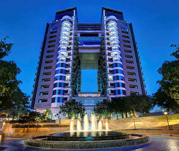 Moderni rakennus, jossa on kaksi tornia yhdistettynä sillalla. Etualalla on suihkulähde puiden ympäröimänä. (Tekoälyn luoma teksti)