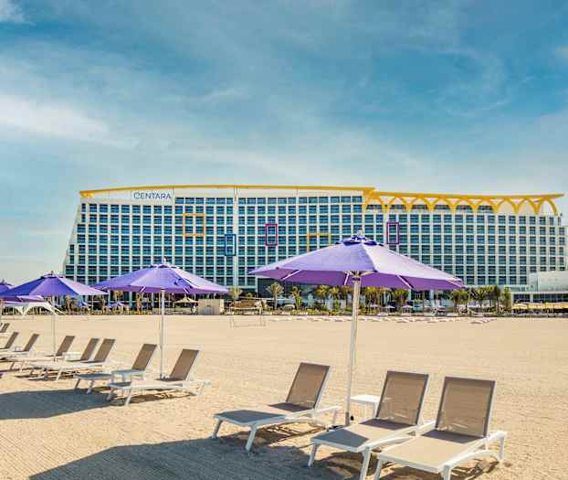 Kuvassa on hiekkaranta aurinkotuoleilla ja violeteilla aurinkovarjoilla. Taustalla näkyy suuri monikerroksinen rakennus. (Tekoälyn luoma teksti)