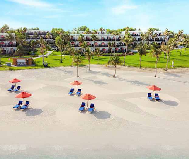 Hiekkaranta, jossa aurinkotuoleja ja aurinkovarjoja, taustalla vihreä alue ja rakennuksia. (Tekoälyn luoma teksti)