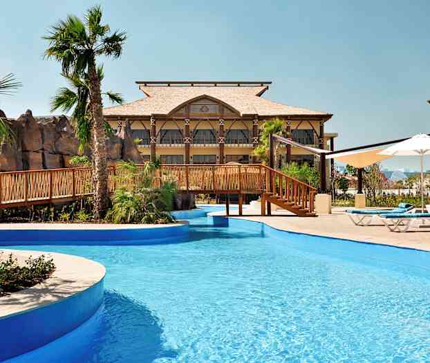 Uima-allas, jossa on sininen vesi ja puinen silta, ympärillä palmuja ja rakennuksia. (Tekoälyn luoma teksti)