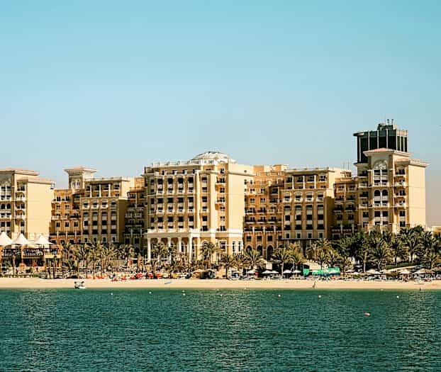 Suuri hotellirakennus rannan äärellä, edessä palmuja, taustalla kirkas taivas ja etualalla meri. (Tekoälyn luoma teksti)