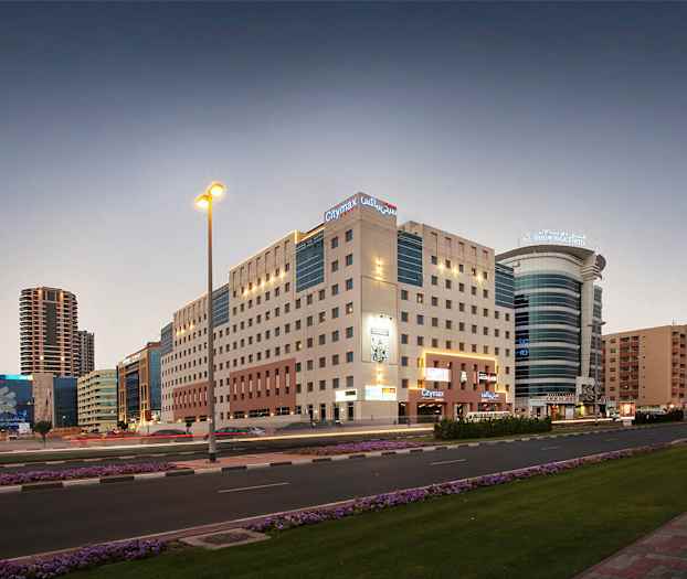 Kaupunkimaisema hämärässä moniasuntoisten rakennusten ja modernien hotellien keskellä. Tie, jolla on liikennettä, ja kukkaistutuksia jalkakäytävän varrella. (Tekoälyn luoma teksti)