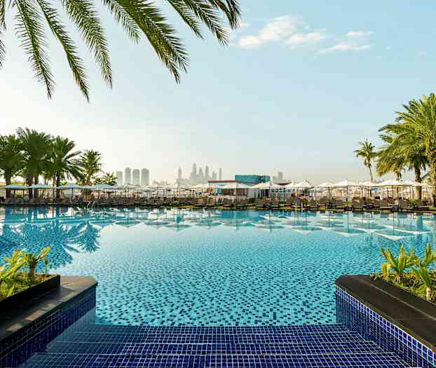 Hotellin uima-allas, jota ympäröivät varjot ja palmut, taustalla kaupunkisiluetti. (Tekoälyn luoma teksti)