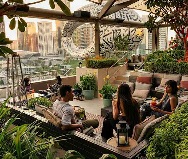 Ulkoilualue, jossa on istumapaikkoja ja vehreyttä ympärillä, kaupunginäkymä ja moderni rakennus taustalla. (Tekoälyn luoma teksti)