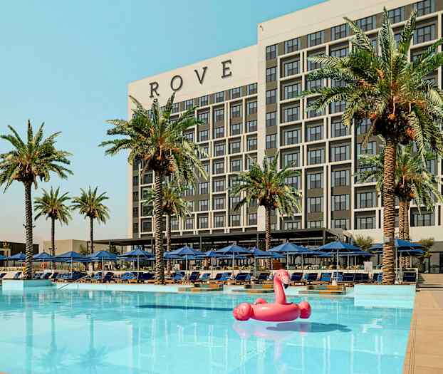 Rakennus, johon on merkitty 'ROVE', uima-altaan takana, ympärillä palmuja ja aurinkotuoleja sekä vaaleanpunainen flamingo-puhallettava lelu vedessä. (Tekoälyn luoma teksti)