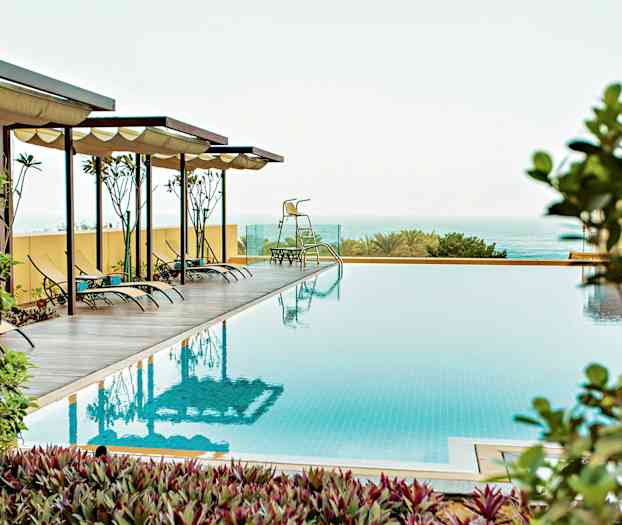 Kuva ulkouima-altaasta, jossa on aurinkotuoleja varjollisten katosten alla, ympärillä kasvillisuutta ja taustalla meri. (Tekoälyn luoma teksti)