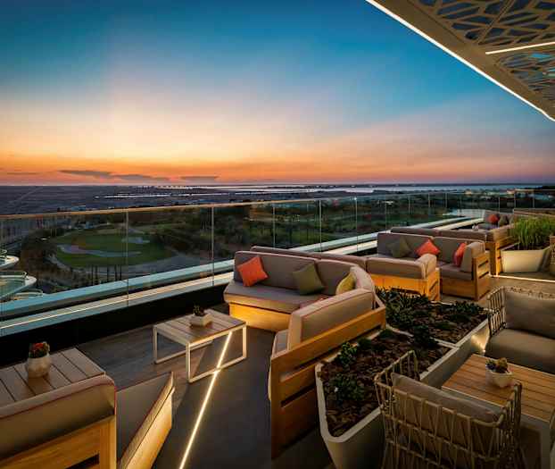 Kattoterassi, jossa on tyylikkäitä istuinryhmiä ja lasikaide, tarjoaa panoraamanäkymän maisemaan auringonlaskun aikaan. (Tekoälyn luoma teksti)