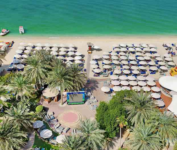 Kuva rannasta, jossa on aurinkovarjoja, aurinkotuoleja, kaksi venettä vedessä sekä palmupuita ja vehreyttä etualalla. (Tekoälyn luoma teksti)