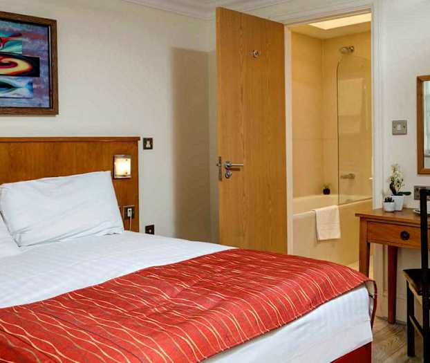 Hotellihuone, jossa sänky, työpöytä, peili ja näkymä kylpyhuoneeseen. (Tekoälyn luoma teksti)