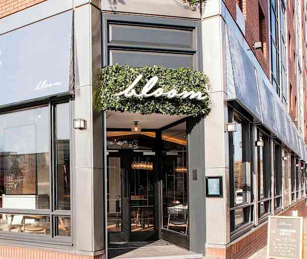 Bloom-ravintolan sisäänkäynti suurilla ikkunoilla ja kyltti, jossa on nimi, ympäröity vihreällä. (Tekoälyn luoma teksti)