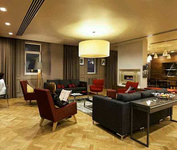 Tyylikäs olohuone, jossa sohvia, nojatuoleja ja sohvapöytä, ympäröitynä lämpimällä valaistuksella ja modernilla sisustuksella. (Tekoälyn luoma teksti)