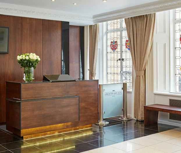 Hotellin vastaanotto, jossa on puinen tiski ja maljakko valkoisia kukkia, koristeellisen ikkunan vieressä. (Tekoälyn luoma teksti)