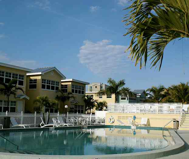 Uima-allas, jota ympäröivät aurinkotuolit, varjot ja talot, trooppiset palmut taustalla. (Tekoälyn luoma teksti)