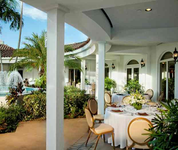 Ulkoterassi, jossa on suihkulähde ja pyöreitä pöytiä, jotka on koristeltu valkoisilla liinoilla ja kukka-asetelmilla. (Tekoälyn luoma teksti)