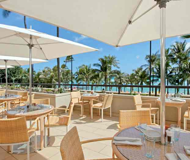 Ulkoterassi, jossa pöytiä ja tuoleja aurinkovarjojen alla, taustalla merinäköala ja palmupuita. (Tekoälyn luoma teksti)