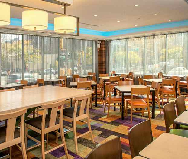 Huone, jossa on paljon pöytiä ja tuoleja, isot ikkunat verhoilla sekä värikkäitä mattoja lattialla. (Tekoälyn luoma teksti)