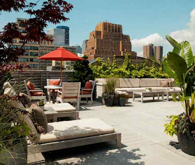 Tilava kattoterassi, jossa ulkokalusteita, aurinkovarjo ja kasveja. Taustalla näkyy rakennuksia ja kirkas taivas. (Tekoälyn luoma teksti)