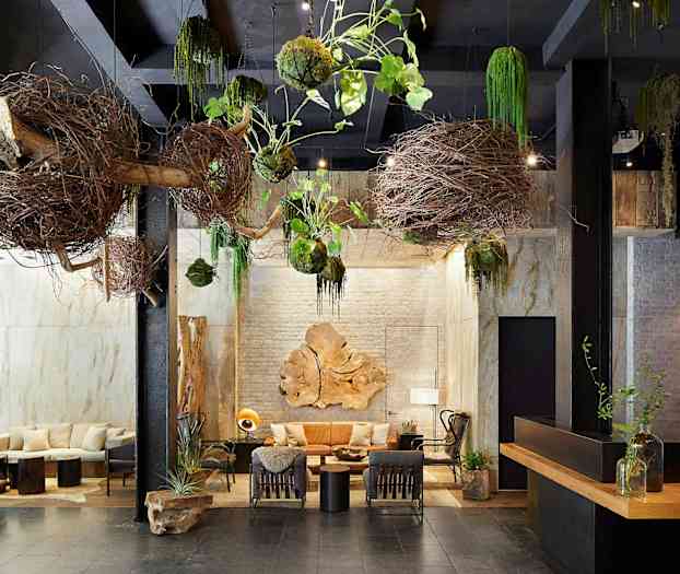 Sisätilan sisustus, jossa on luonnollisia koristeita, kuten oksia ja kasveja, jotka riippuvat katosta. Keskellä on istuimia ja pöytiä koristeluineen. (Tekoälyn luoma teksti)