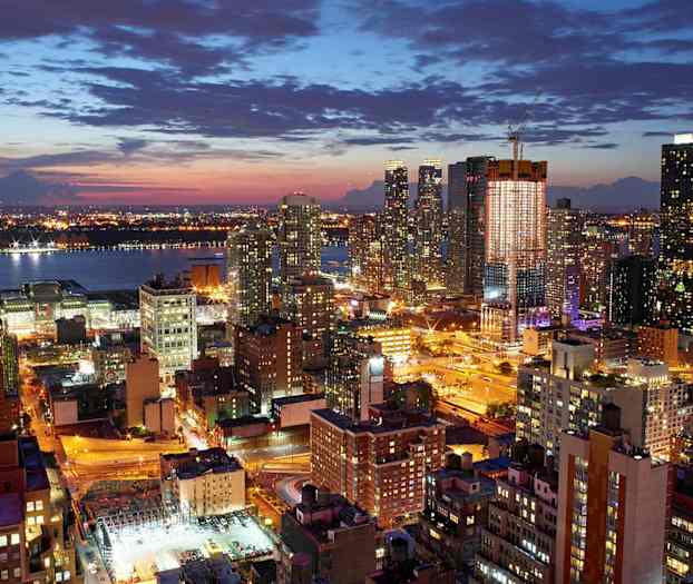 Yöaikainen kaupunkimaisema, jossa on useita valaistuja rakennuksia ja katuja, sekä joki taustalla. (Tekoälyn luoma teksti)