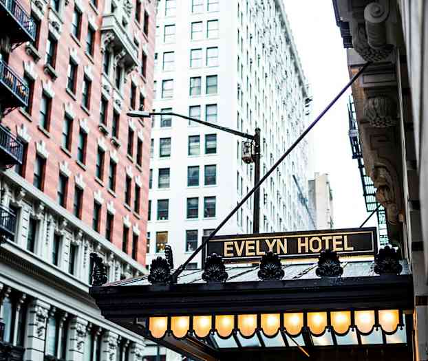 Kuvassa on Evelyn-hotellin sisäänkäynti valaistulla kyltillä ja ympäröivien rakennusten julkisivut. (Tekoälyn luoma teksti)