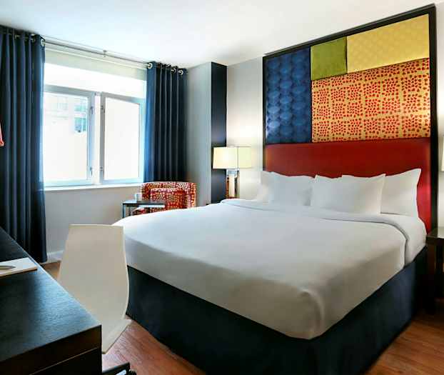 Hotellihuone, jossa iso sänky, tummat verhot, työpöytä ja värikäs seinäkoriste. (Tekoälyn luoma teksti)