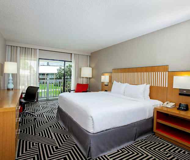 Hotellihuone, jossa on suuri vuode, työpöytä ja tuoli sekä parveke, jolta on näkymä nurmikolle. (Tekoälyn luoma teksti)